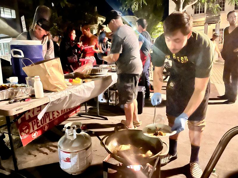 A man wearing gloves cooks food in a large pan outdoors at night, while others prepare and serve food at tables in the background.