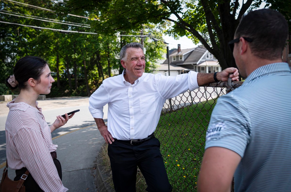 A man in a white shirt leans on a chain-link fence, engaging in conversation with a woman holding a smartphone and another man in a striped shirt, outdoors on a sunny day.