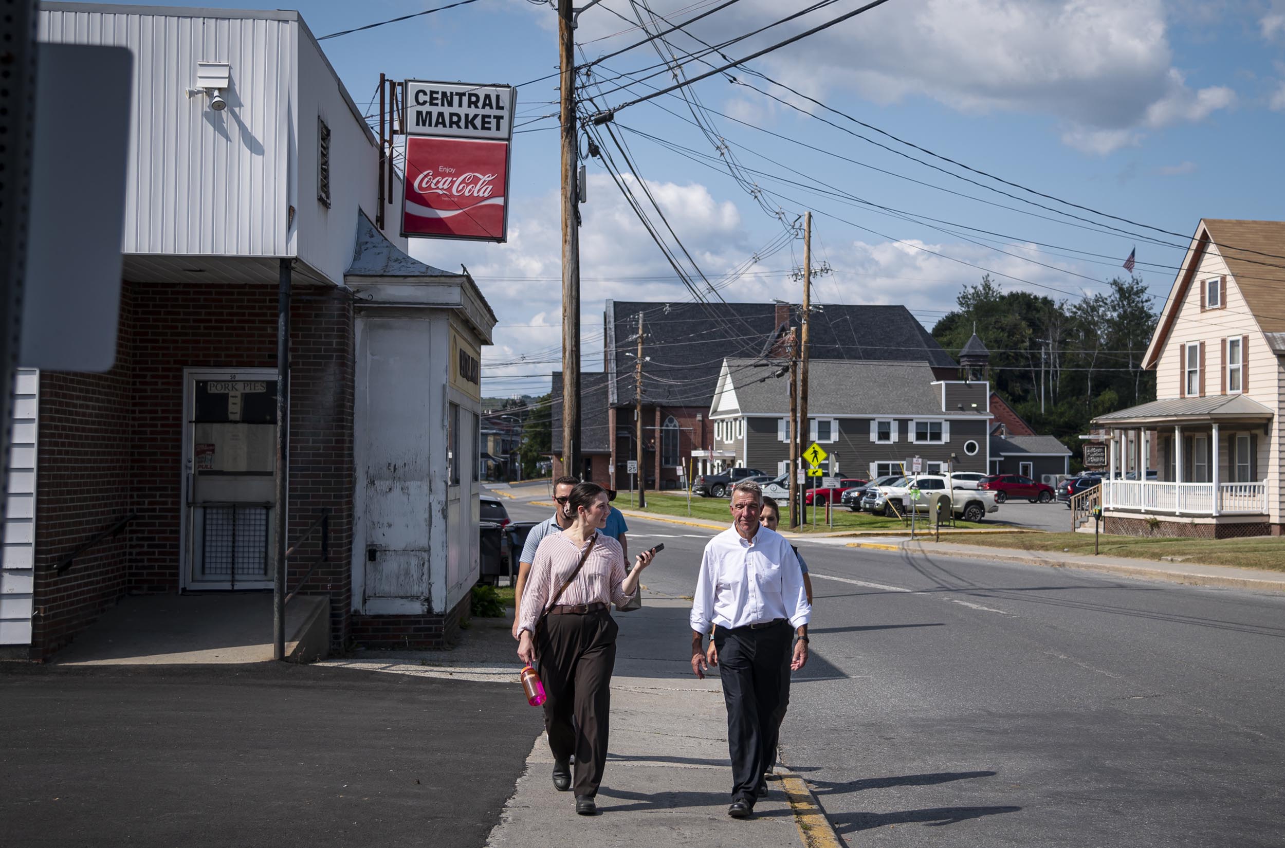 Two people walk along a sidewalk next to a Central Market store. There are houses and power lines in the background under a partly cloudy sky.