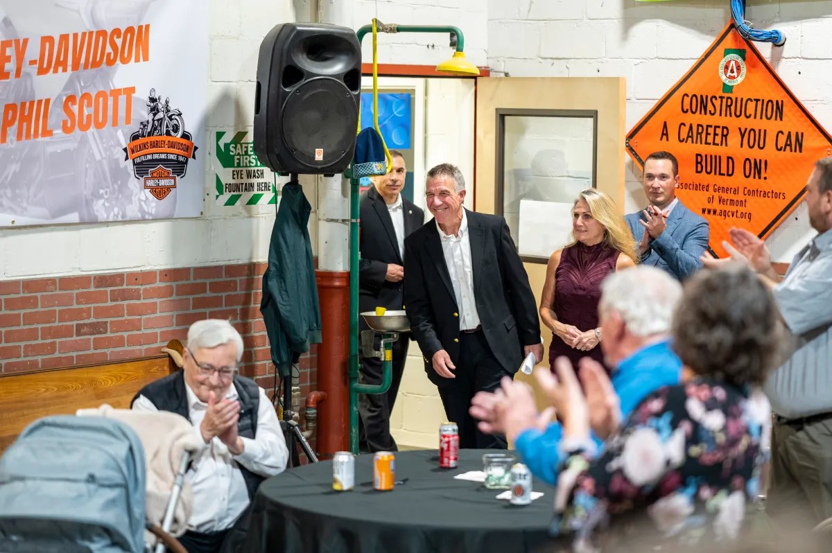 A group of people in a room, some seated and others standing near a Harley-Davidson banner. A man in a suit walks in, greeted by applause. A construction sign is visible on the wall.
