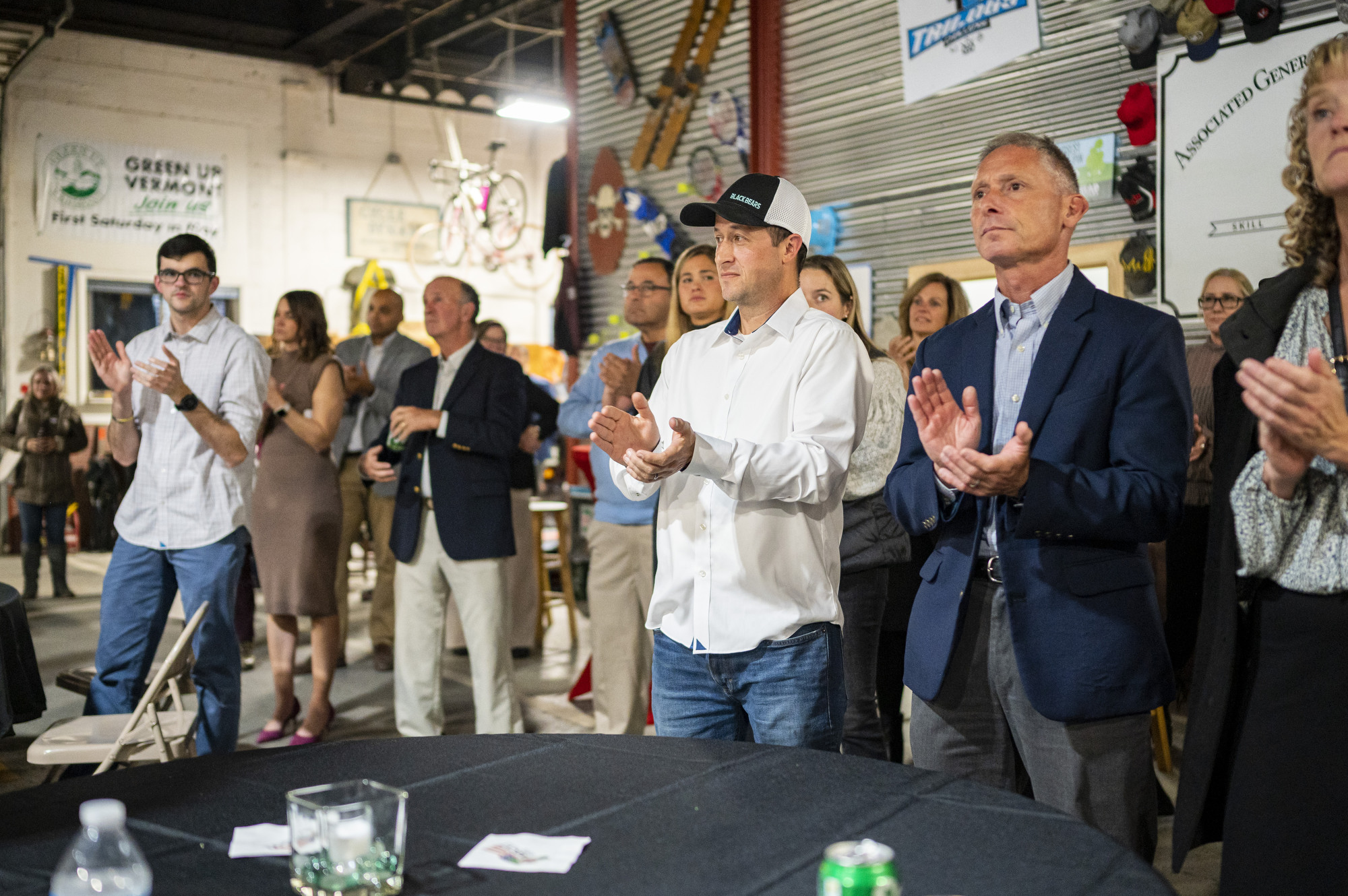 A group of people stands and applauds in a casual indoor setting with posters and bicycles on the walls.