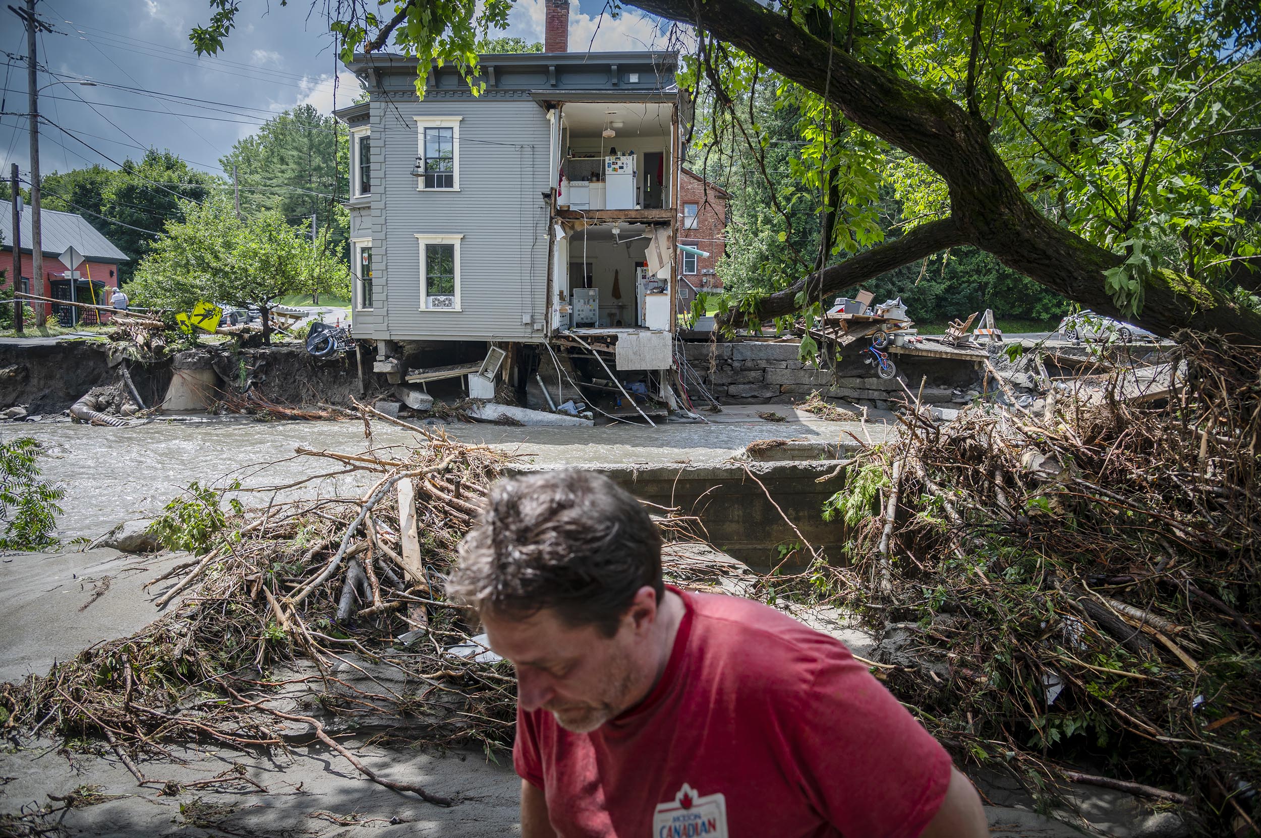 A man in a red shirt stands in front of a damaged house partially collapsed by a flood, with debris and uprooted trees surrounding the area.