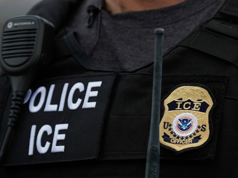 Close-up of a person wearing a vest labeled "POLICE ICE" with an ICE officer badge and a Motorola radio microphone attached.