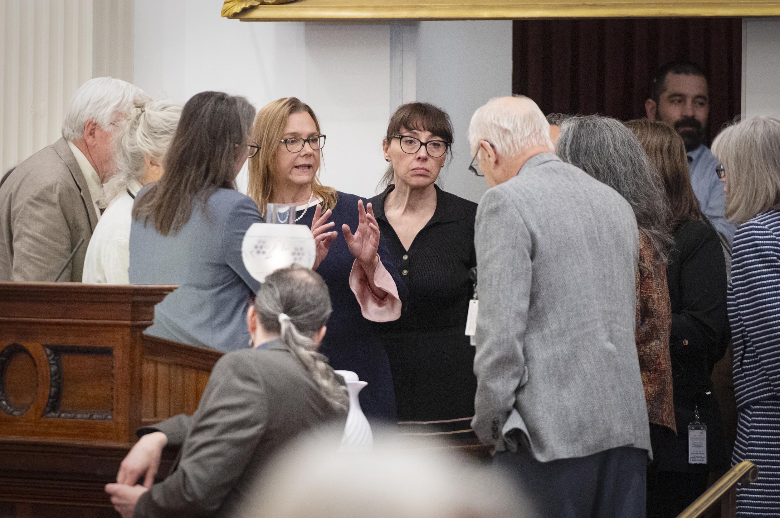 A group of people stand closely together in discussion near a wooden podium in a formal indoor setting.