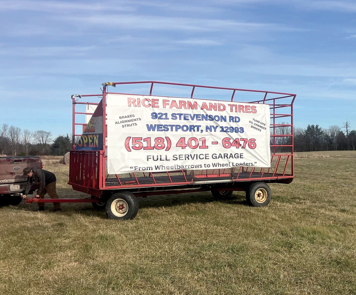 A person attaches a red trailer with a sign for "Rice Farm and Tires" to a vehicle in a grassy field. The sign displays contact details and services offered.