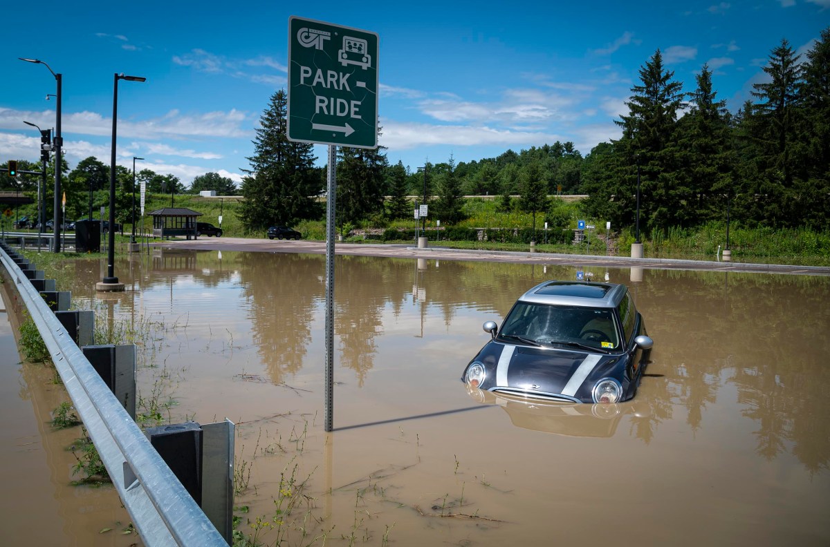 A car partially submerged in floodwater in a parking lot with a "Park and Ride" sign visible.