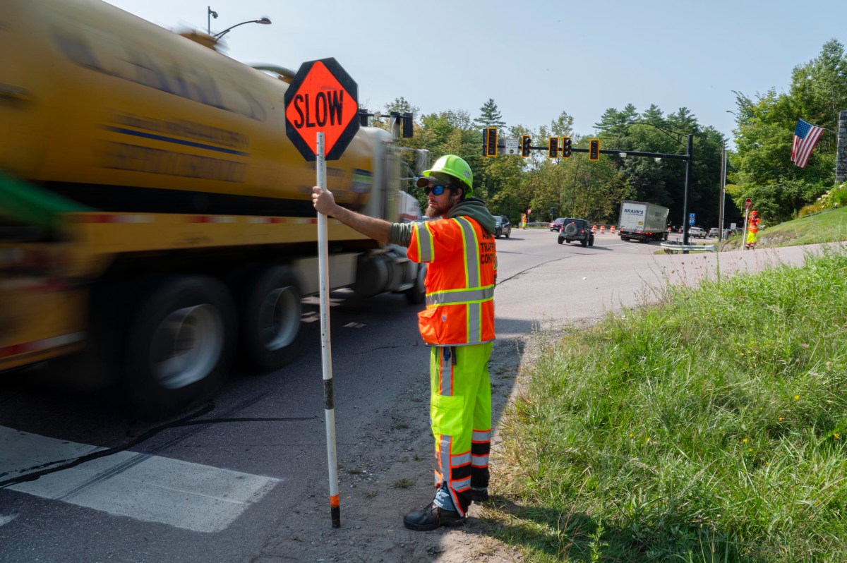 A construction worker in a safety vest and helmet holds a "SLOW" sign as traffic, including a large truck, passes by on a road near a grassy area.