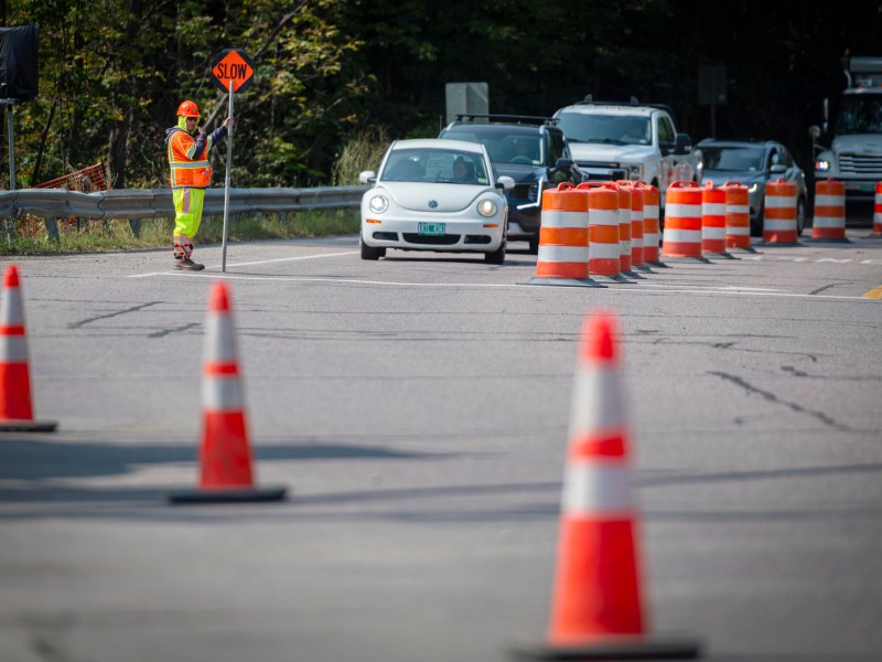 A construction worker in a safety vest holds a "SLOW" sign as cars wait in a lane marked by orange traffic barrels and cones on a road.