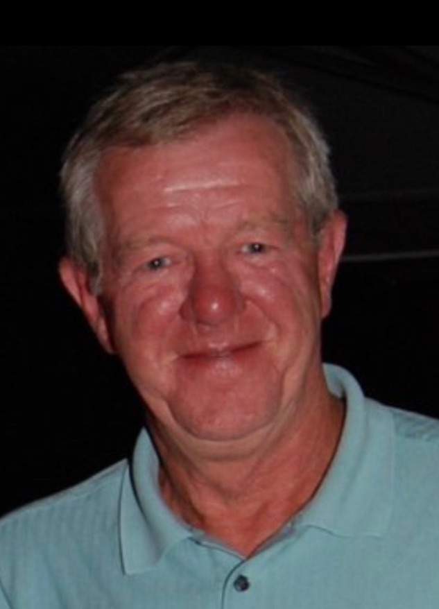 A man with short hair and a light blue polo shirt smiles at the camera against a dark background.