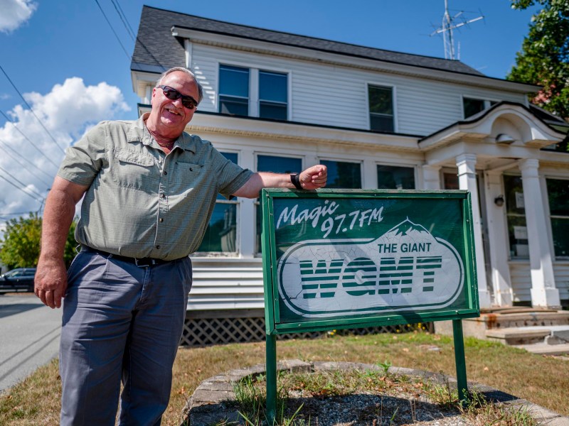 A man wearing sunglasses stands next to a sign for radio station Magic 97.7 FM WGMT in front of a white house on a sunny day.