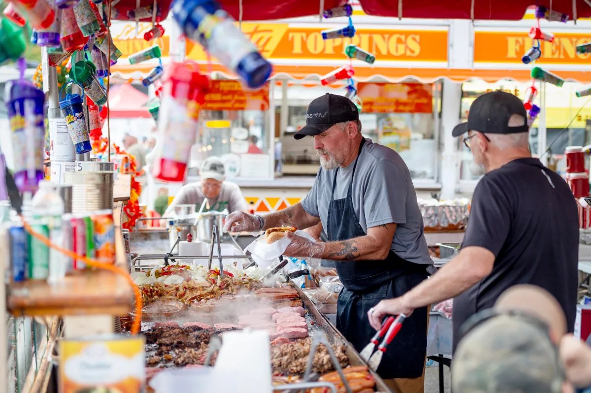 Two men cook and assemble food at a busy outdoor market stall, surrounded by colorful decorations and various meats grilling on an open barbecue.