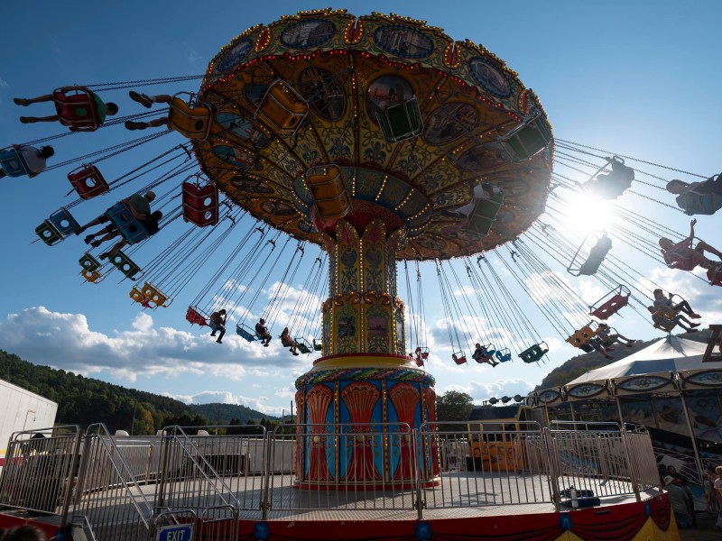 A brightly colored swing ride spins with people in seats at an outdoor amusement park, with sunlight and blue sky in the background.