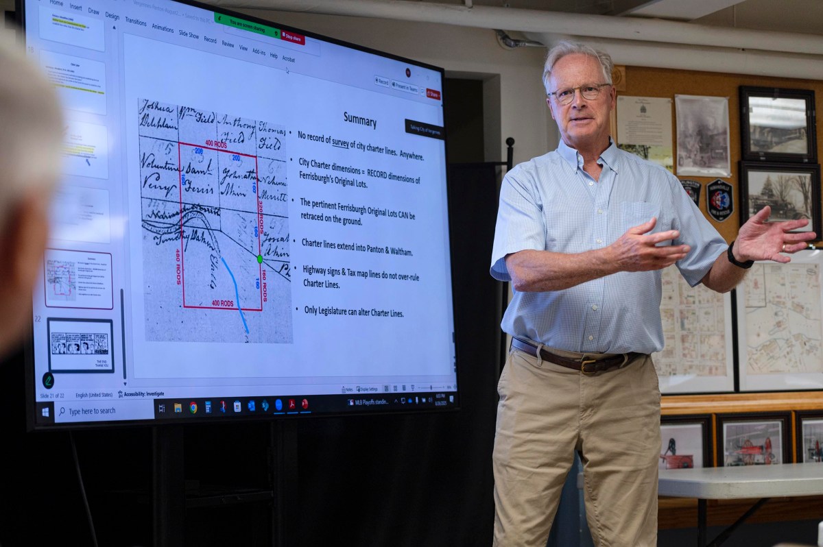 A man stands next to a large screen displaying a map and summary text, giving a presentation in a classroom or meeting room.
