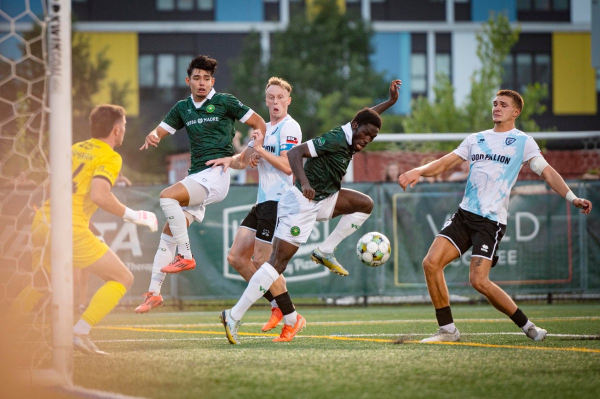 Several soccer players in green and white jerseys compete for the ball near the goal as a goalkeeper watches, with a modern building in the background.