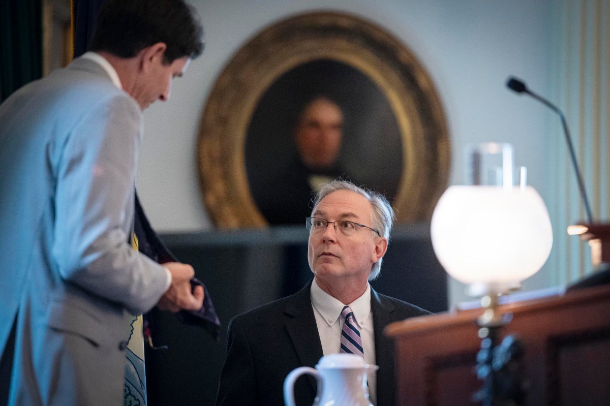 Two men in suits are engaged in conversation in a formal room. One is standing and holding a tie, and the other is seated and looking up. A portrait hangs in the background.