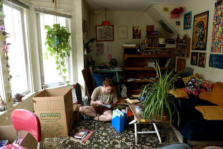 A person sits on the floor of a cluttered living room, surrounded by boxes, plants, books, and eclectic decor, appearing to work or read.