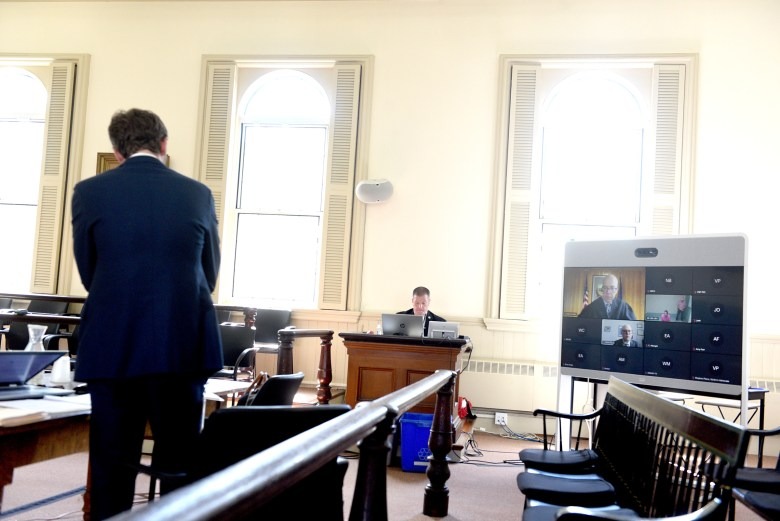 A person stands in a courtroom while another sits at a desk; a large screen displays participants joining the court proceedings via video call.