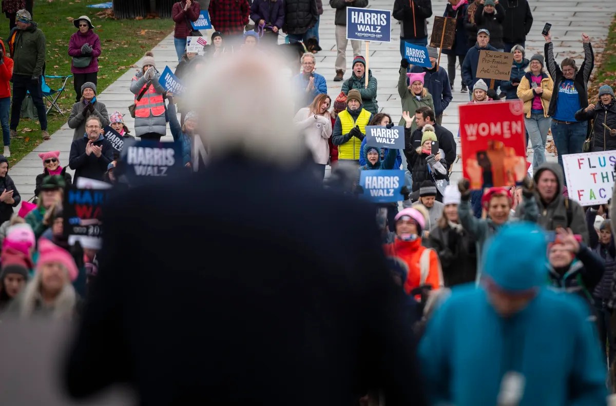 A crowd gathers at a rally with people holding signs, some reading "Harris Walz" and "Women." A speaker is in the foreground, blurred.