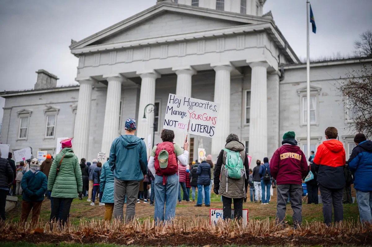 People gathered in front of a courthouse holding signs, one reads, "Hey Hypocrites, Manage Your Guns, Not My Uterus.