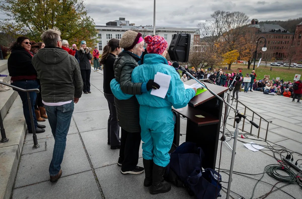 A person speaks at a podium with an arm around another person in a blue outfit. A crowd is gathered nearby, listening.