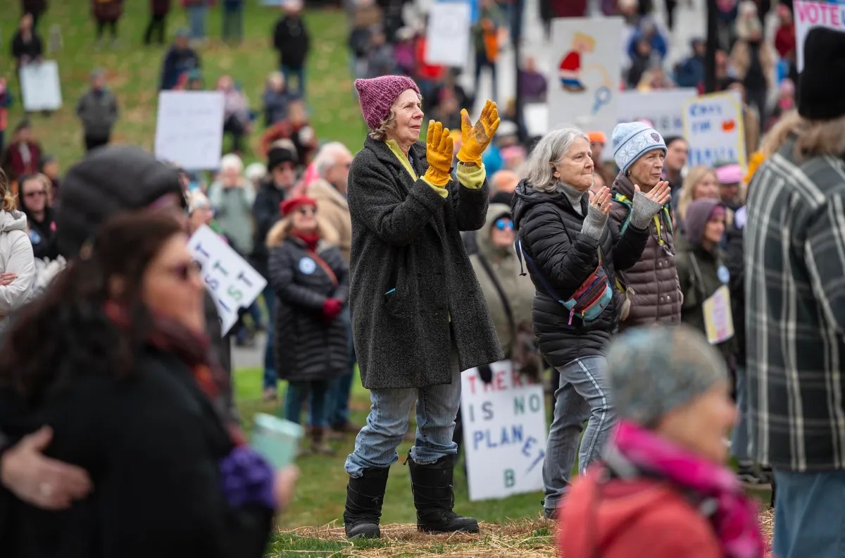 A crowd of people at an outdoor rally, with some holding signs. Central figures include a person clapping in a purple hat and yellow gloves, and another standing beside them.