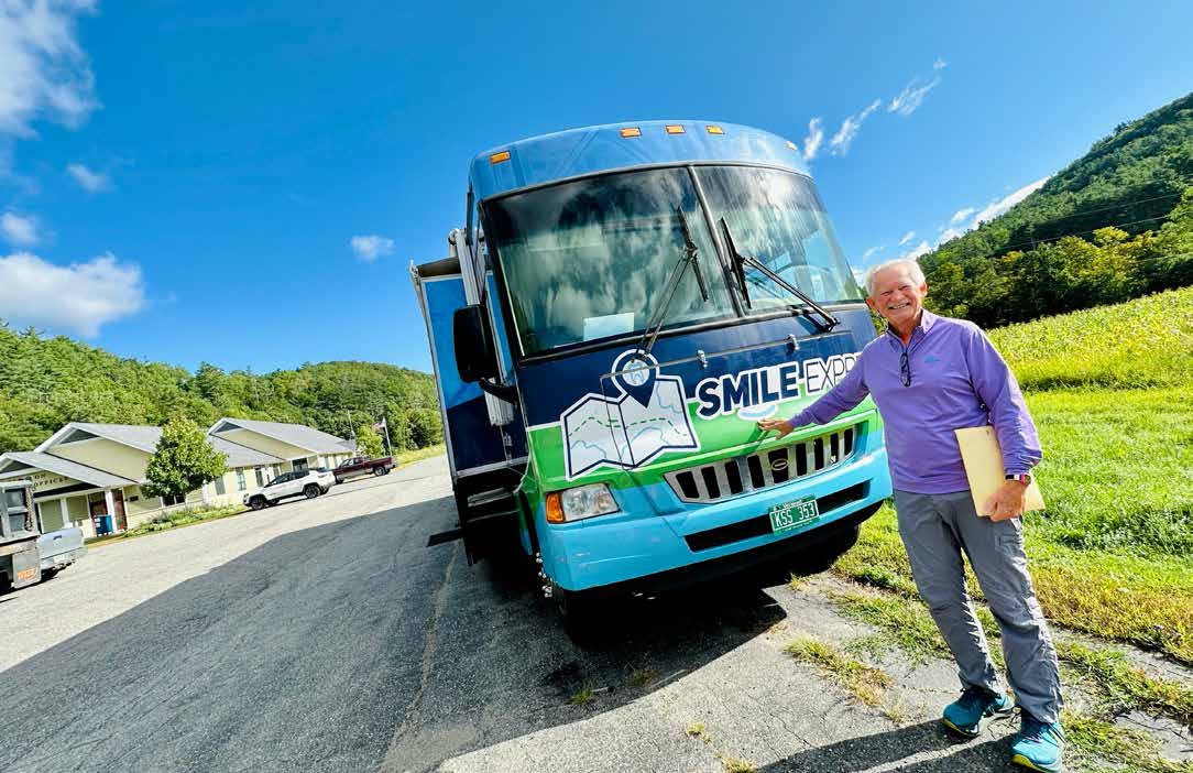 A man stands next to a blue truck labeled "Smile Express" on a sunny day, holding a folder, with houses and green hills in the background.